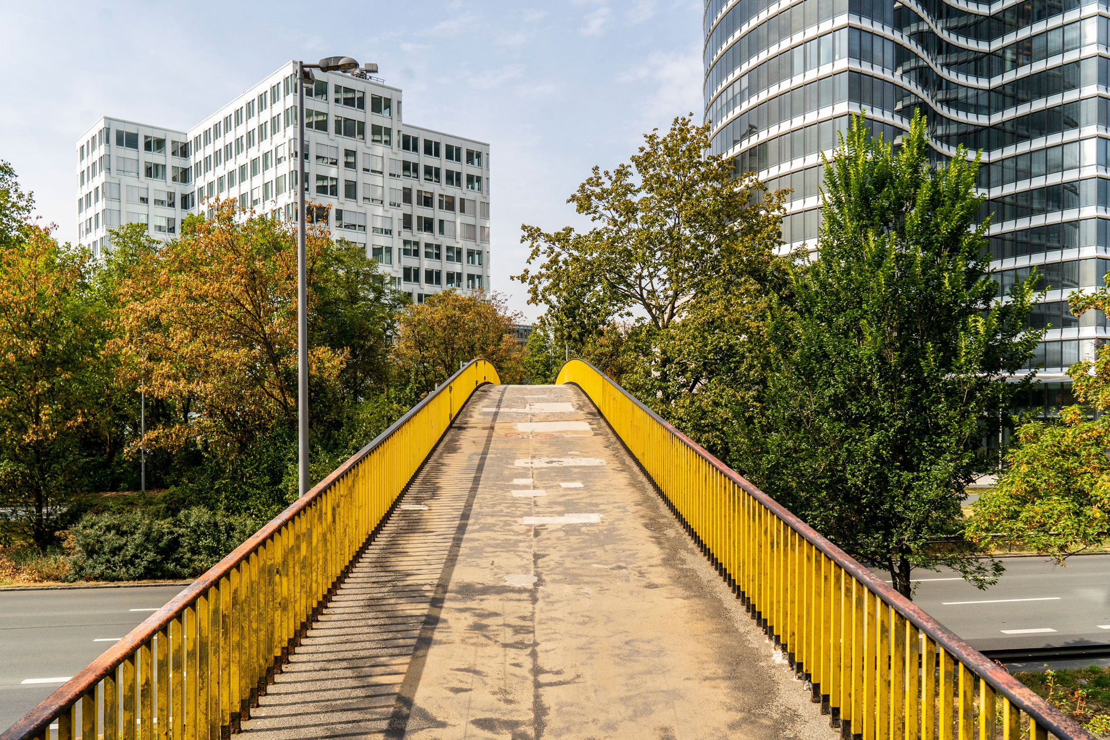 View of a bridge over a multi-lane road. The railing is painted yellow and trees have been planted next to the bridge. The bridge runs towards several high-rise buildings.