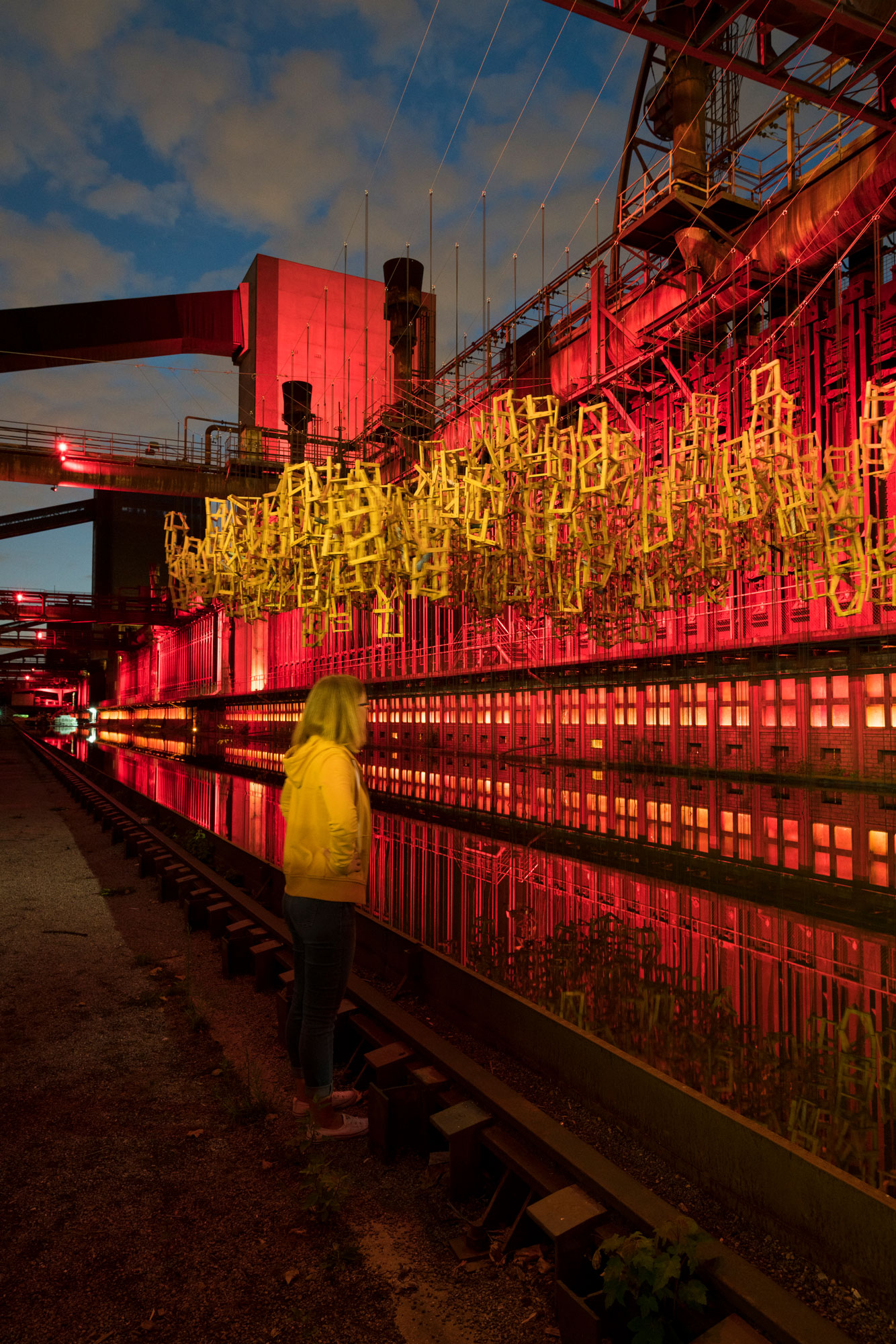 A woman stands in front of a red-lit industrial building at night.