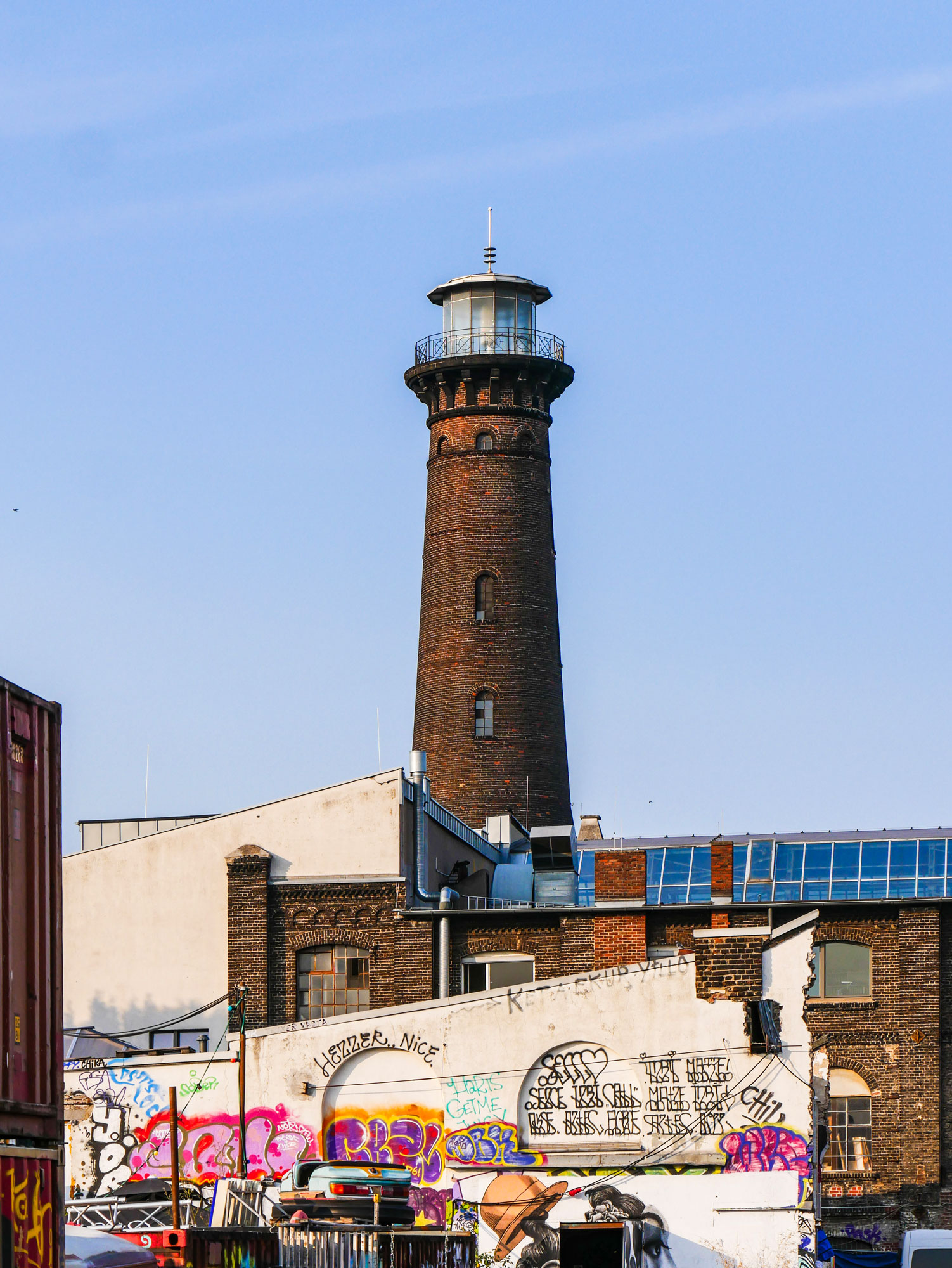 View of a lighthouse in the middle of Cologne. In front of it you can see half-dilapidated houses and walls that are full of graffiti.