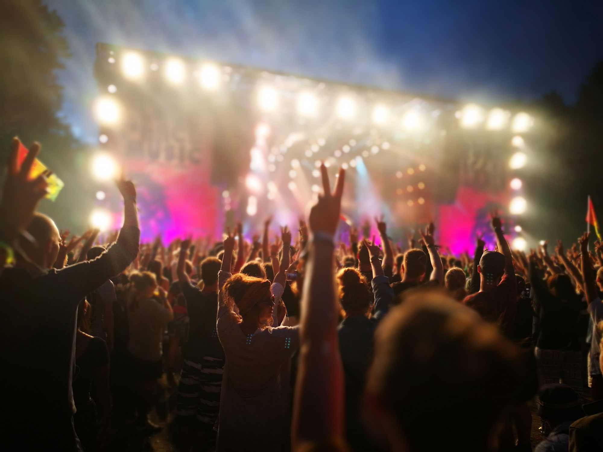 Ansicht einer beleuchteten Bühne bei Nacht, auf der eine Band spielt. Davor steht eine Zuschauermenge und macht das Peace-Zeichen.