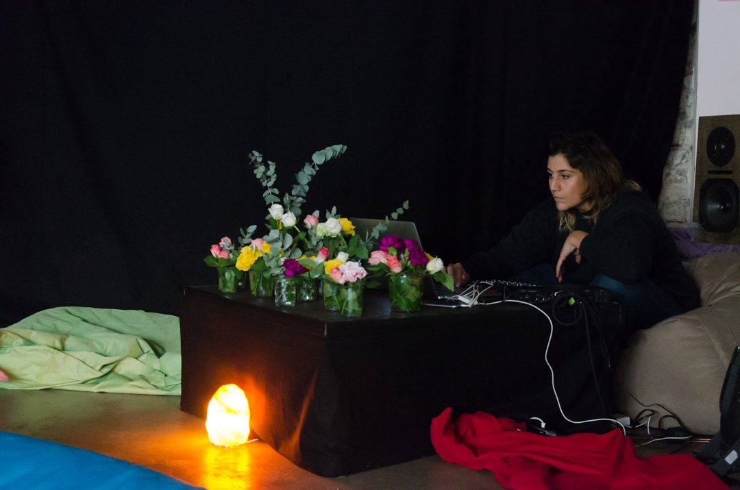 {:gb}Photo of the artist, producer and DJ Neda Sanai. She looks at a laptop in front of which there are many small vases of flowers.{:}{:de}Foto der Künstlerin, Produzentin und DJ Neda Sanai. Sie schaut auf einen Laptop vor dem viele kleine Vasen mit Blumen stehen.{:}